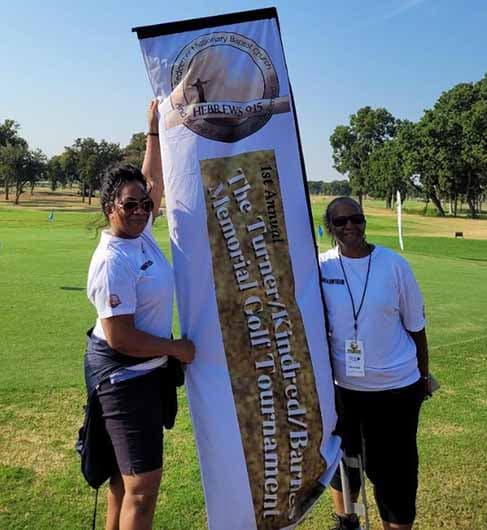Two volunteers hold the 1st Annual Turner/Kindred/Barnes Memorial Golf Tournament banner on the course